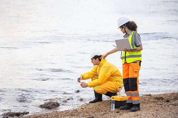 Team of Scientists and Environmental Researchers in Protective Gear Collecting Water Samples on Coastline, Specialized Site Inspectors in Safety Uniforms Documenting Marine Pollution with Laptop